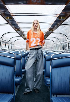 Jong persoon met lang blond haar, gekleed in een oranje trui en brede grijze jeans, staand in een bus met blauwe stoelen en een glazen dak.