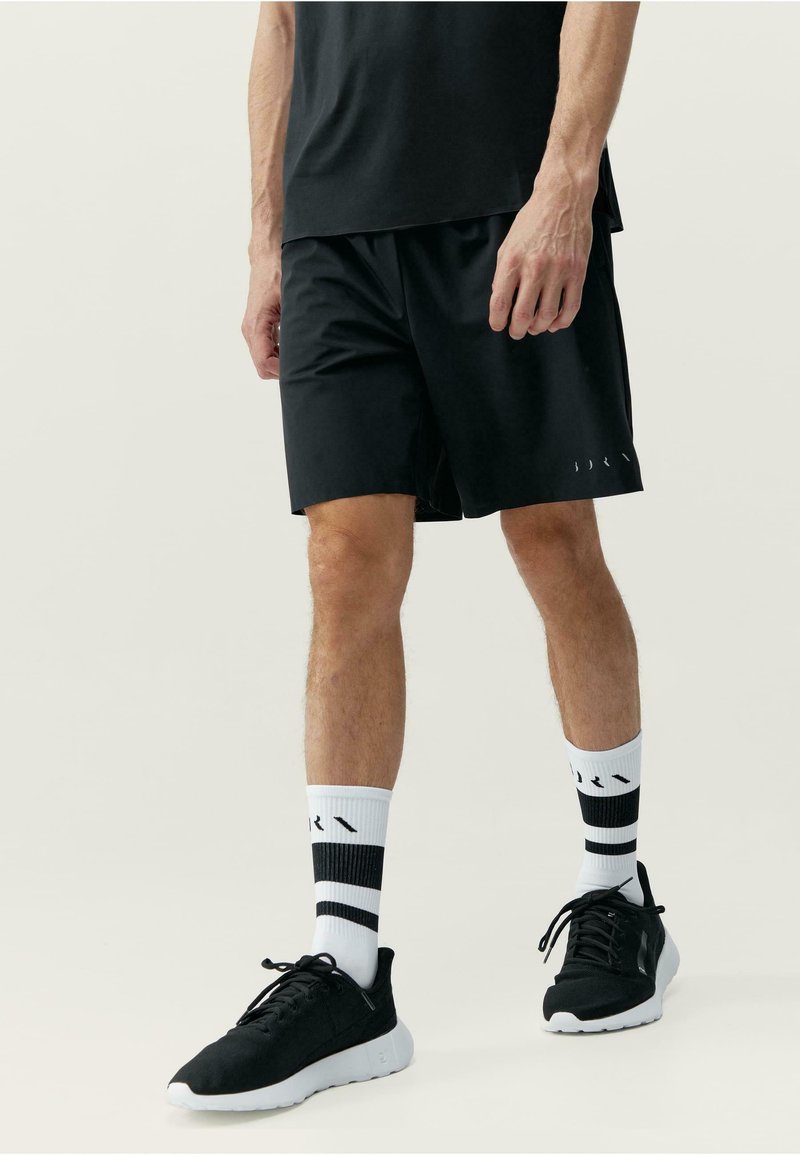 Fekete sportshort, könnyű anyagból készült, laza szabással, finom logórészlettel az alján, feketére passzoló sportcipőkkel és csíkos zoknikkal.