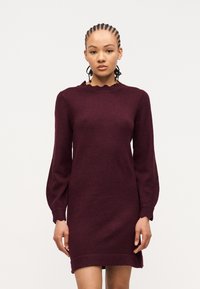 Γυναίκα με πλεξούδες, φοράει μακρυμάνικο μπορντό πλεκτό φόρεμα με κυματιστό ντεκολτέ και μανσέτες, στέκεται μπροστά σε απλό ανοιχτό φόντο.