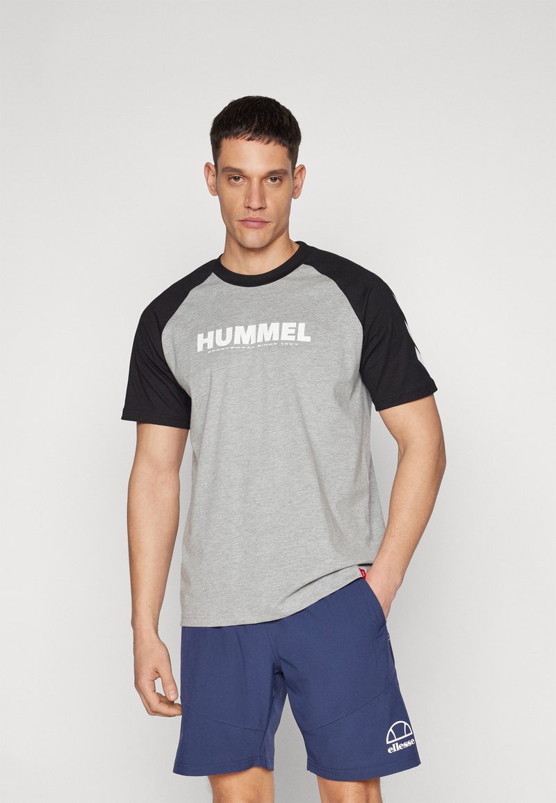Grå T-skjorte med sorte raglanermer, med ordet "HUMMEL" i hvitt på brystet. Satt sammen med marineblå shorts, som viser et enkelt design.