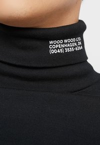 Černý rolák s ribbovanou texturou a vysokým límcem. Na něm je bílé písmo: „WOOD WOOD LTD. KODAŇ, DK (0045) 3535-6264.“