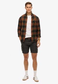 Geruite flanellen shirt in zwart, oranje en groen; witte T-shirt; grijze cargoshort; witte en zwarte sneakers; beige sokken.