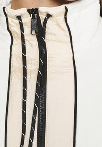 Veste à fermeture éclair blanche avec un col montant, présentant un panneau avant beige et des accents noirs. Tissu texturé et cordons contrastants.