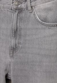 Gros plan sur le devant d'un jean en denim gris avec un bouton en métal, une passants de ceinture et les coutures de la poche avant visibles.