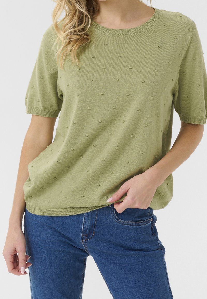 Femme portant un haut en maille vert clair à manches courtes avec de petits points texturés et un jean en denim bleu, posant avec une main dans la poche.