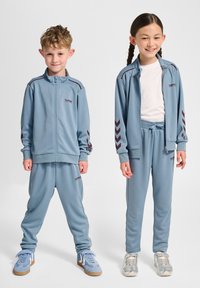 Niebieskie dresy z czerwonymi akcentami, z kurtką na zamek i spodniami z elastycznym pasem, wykonane z gładkiego materiału. W zestawie dziecięce obuwie sportowe.