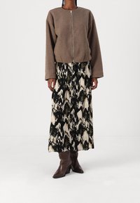 Žena v hnědém fleecovém zipovém svršku, s černobílou abstrahující midi sukní a tmavě hnědými kozačkami na podpatku, stojící na bílé podlaze.