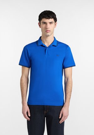 Giovane uomo con capelli corti e ricci indossa una polo blu con rifiniture nere su colletto e maniche, in piedi davanti a uno sfondo bianco.