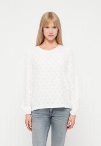 Witte blouse met lange mouwen, voorzien van een gatenpatroon, ronde halslijn en geplooide manchetten. Gepaard met lichtblauwe jeans.
