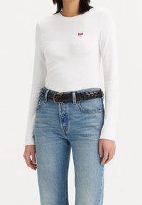 Valkoinen pitkähihainen paita, jossa on pieni punainen Levi's-logotakki rinnan kohdalla, yhdistettynä vaaleansinisiin kulutettuihin suoriin farkkuihin ja mustaan punottuun vyöhön.