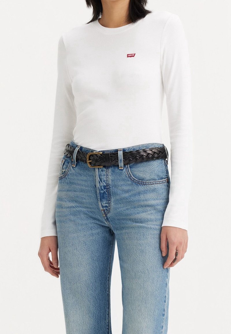 Valkoinen pitkähihainen paita, jossa on pieni punainen Levi's-logotakki rinnan kohdalla, yhdistettynä vaaleansinisiin kulutettuihin suoriin farkkuihin ja mustaan punottuun vyöhön.