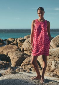 Femme portant une robe rose sans manches ornée de motifs de coquillages et des tongs, debout sur une rive rocheuse avec l'océan et un ciel clair en arrière-plan.