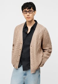 Νέος άνδρας με γυαλιά, που φοράει μπεζ πλεκτή ζακέτα με σχέδιο κοτσίδα πάνω από μαύρο πουκάμισο και ανοιχτόχρωμα μπλε τζιν, στέκεται με τα χέρια στις τσέπες.