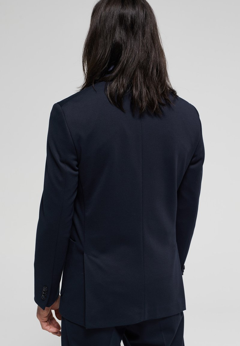 Chaqueta de traje azul marino con un corte entallado, abertura trasera y detalles de puños abotonados. Textura de tela suave y diseño estructurado.