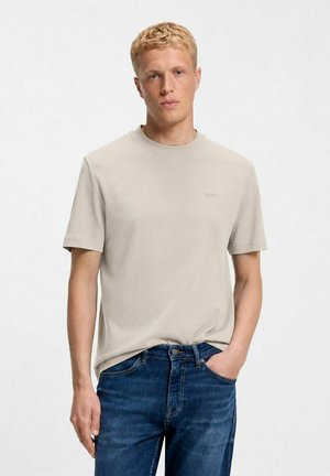 Man met kort blond haar, die een beige shirt met korte mouwen en blauwe spijkerbroek draagt, staat met één hand in zijn broekzak tegen een effen achtergrond.