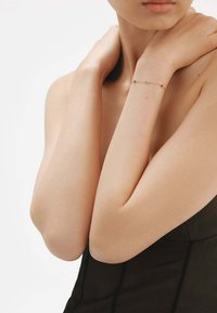 Delicada pulsera de oro con pequeñas piedras preciosas de colores múltiples dispuestas a intervalos, llevada en una muñeca desnuda. Piel suave y ropa mínima visible.