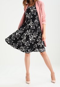 Robe noire avec motif floral blanc, silhouette en A, associée à un cardigan rose clair et des talons beiges. Tissu lisse, longueur cheville.