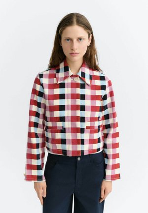 Mujer con chaqueta corta a cuadros roja, blanca, azul y negra con un gran cuello y pantalones oscuros sobre un fondo blanco.