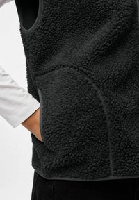 Melna vilnas veste ar teksturētu, pūkainu virsmu, sānu kabatām un pelēku apmali gar malām. Rokas paliek kabatā.