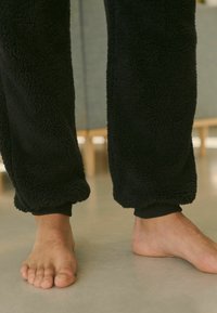 Fekete, bolyhos fleece joggingnadrág, elasztikus mandzsettával, mezítláb egy sima padlón. Az anyag lágy és textúrált.
