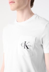 Zblízka osoba v bílém tričku s kapsou, na které je logo "ck Calvin Klein Jeans" na hrudi.