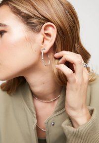 Boucle d'oreille en argent avec une finition textur�ée, de forme ovale et un design minimaliste, complétée par une veste bomber de couleur neutre.
