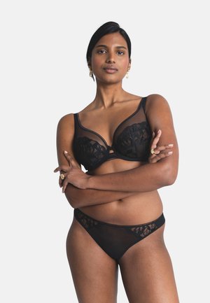 Mujer de pie con los brazos cruzados, vestida con un conjunto de lencería negro de encaje, sujetador y bragas a juego, sobre un fondo liso.