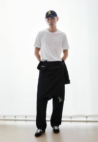 Witte korte mouwen T-shirt, donkere denim wikkelrok, brede donkere broek, zwarte sneakers met witte veters en een marineblauwe pet met een geel logo.