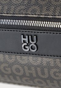 Material preto texturizado com um fecho éclair prateado e o logótipo "HUGO" em relevo, realçado por uma tira de pele preta suave acima do fecho éclair.