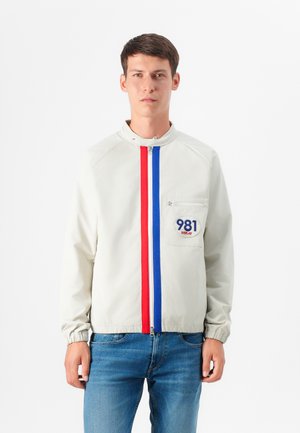 Un jeune homme se tient contre un fond blanc, portant une veste claire avec des rayures rouges et bleues ainsi que des jeans, regardant directement le spectateur.