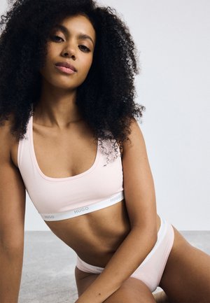 Femme aux cheveux bouclés portant un soutien-gorge de sport et des sous-vêtements Hugo de couleur rose clair, assise sur le sol contre un fond uni.