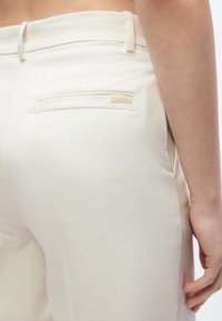 Κοντινό πλάνο σε παντελόνι κρεμ χρώματος με πίσω τσέπη welt και θηλιές για ζώνη, φορεμένο από άτομο που έχει το χέρι του ακουμπισμένο δίπλα στο σώμα.