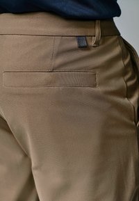 Braune Hosen mit glatter Textur, versehen mit einer Gesäßtasche mit einem dezenten horizontalen Design und einem kontrastierenden Stoffschlaufe.