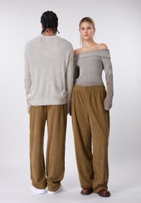 Δύο φιγούρες στέκονται πλάτη με πλάτη. Η μία φοράει ανοιχτό γκρι πλεκτό πουλόβερ και αντιστοιχούντα χαλαρά παντελόνια από κορδόνι· η άλλη φοράει γκρι τοπ με ακάλυπτους ώμους και καφέ παντελόνια.