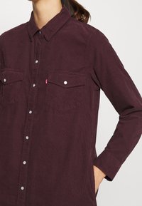 Chemise en velours côtelé bordeaux à manches longues, fermeture avant à boutons, deux poches poitrine, et boutons-pression blancs. Présente une texture côtelée distincte.