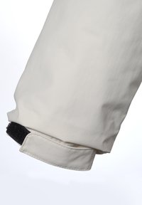 Lys beige jakkeærme, tæt på, med en glat tekstur, rund manchet og en sort Velcro-rem til justering.