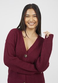 Młoda kobieta z długimi ciemnymi włosami, uśmiechająca się, ubrana w bordowy sweter z dużymi guzikami, pozująca na jednolitym jasnym tle.