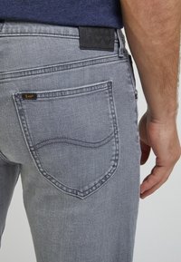 Man wearing light gray Lee jeans with visible back pocket and hand resting beside thigh against plain background.