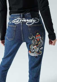 Tummansininen farkkuhousut, joissa on brodeerattu pääkallo, käärme ja kotka -kuvioita. "Ed Hardy" -logo on selvästi ommeltu taakse.