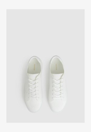 Valged spordijalatsid, mis on valmistatud struktuursetest kudumismaterjalidest, tugevdatud paeltega, ümarate varvastega ja diskreetse logoga keelel.