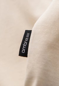 Tejido beige claro con una textura suave, que presenta una etiqueta negra con texto blanco que dice "ombre" parcialmente visible en la esquina.