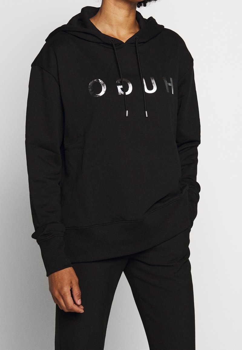 Personne portant un sweat à capuche noir avec des lettres argentées brillantes "HUGO" imprimées à l'envers sur le devant, accompagné d'un pantalon noir.
