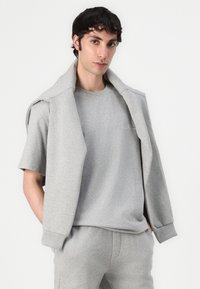 Tricou din bumbac gri cu mâneci scurte, având un guler rotund. O jachetă gri asortată este aruncată peste umeri.
