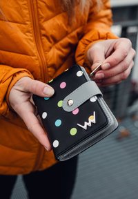 Černá peněženka s barevnými puntíky (růžová, zelená, modrá, bílá) a šedým popruhem, s kovovým logem. Kompaktní obdélníkový tvar.