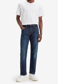 Vaqueros de denim azul oscuro con corte recto, con un bolsillo frontal y costuras en contraste. Combinados con una camiseta blanca lisa y zapatillas blancas.
