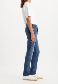 Personne portant un jean droit bleu, un t-shirt blanc et des baskets blanches, se tenant de profil contre un fond blanc.