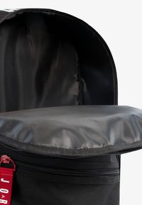 Interior de mochila negra con forro de tela suave, un compartimento acolchado central y un cierre con cremallera. Etiqueta roja con el nombre de la marca visible.