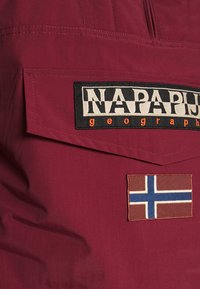 Giacca bordeaux con un patch logo "NAPAPIJRI" in bianco e nero e un patch bandiera con una croce blu e bianca su sfondo marrone.