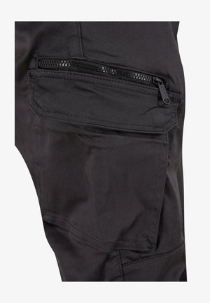 Musta cargo-tasku, jossa on vetoketjullinen suljenta, näkyvillä ompeleilla ja sileä kangaspinta. Tasku on integroitu housun lahkeeseen.