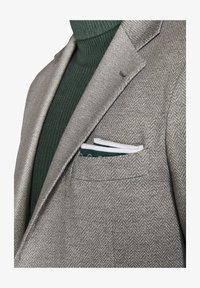 Blazer gris de espiga con solapas de muesca, combinado con un cuello de tortuga acanalado en verde oscuro. Paño de bolsillo en blanco y verde visible en el bolsillo del pecho.